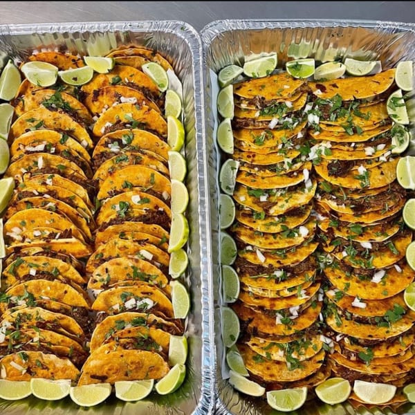 Two foil trays filled with rows of birria tacos, garnished with cilantro, onions, and lime wedges.