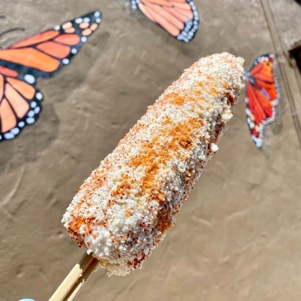 Elote, Mexican street corn, covered in white cheese and red spices, against a butterfly mural.