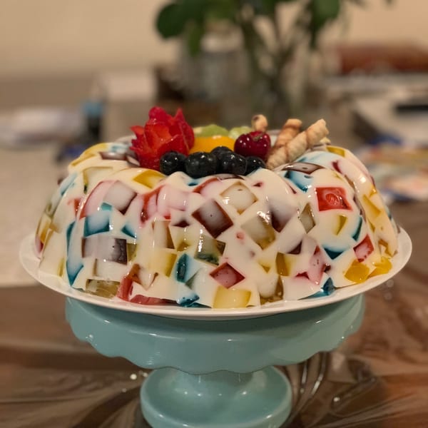 Colorful mosaic gelatin dessert topped with fresh berries, carved strawberry, and wafer rolls on a blue stand.