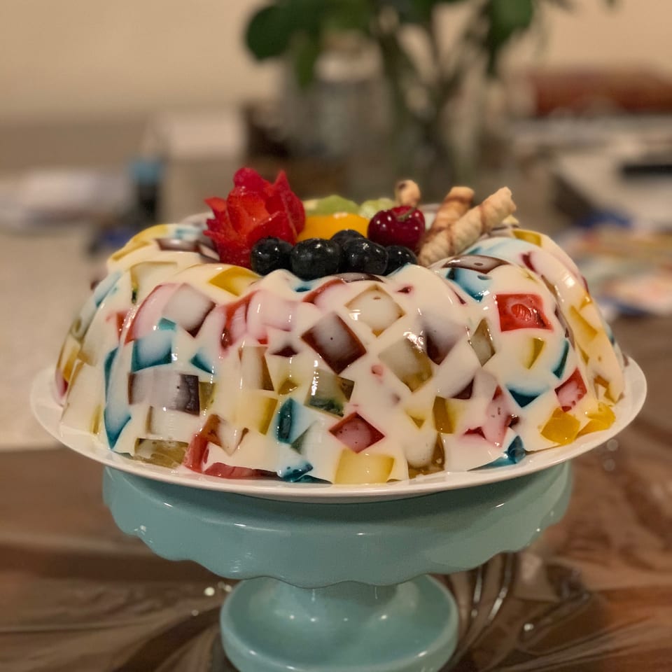 Colorful mosaic gelatin dessert topped with fresh berries, carved strawberry, and wafer rolls on a blue stand.