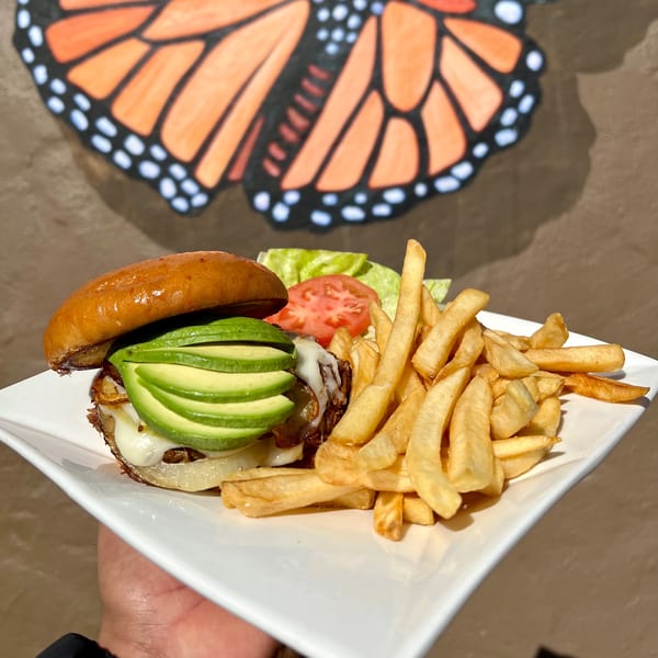 Aquino's Birrieria & Brunch's avocado burger with fries, lettuce, and tomato, against a butterfly mural.