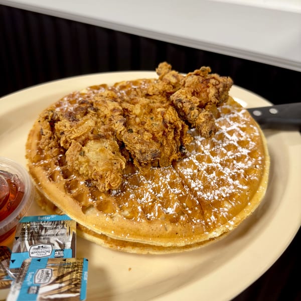 Chicken and waffles with powdered sugar, syrup, and butter packets on a light yellow plate.