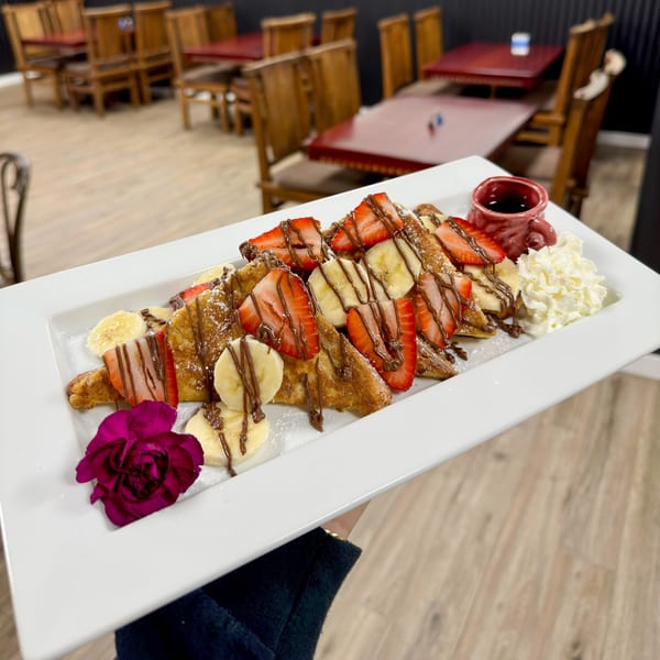French toast with strawberries, bananas, chocolate drizzle, whipped cream, and a purple flower.