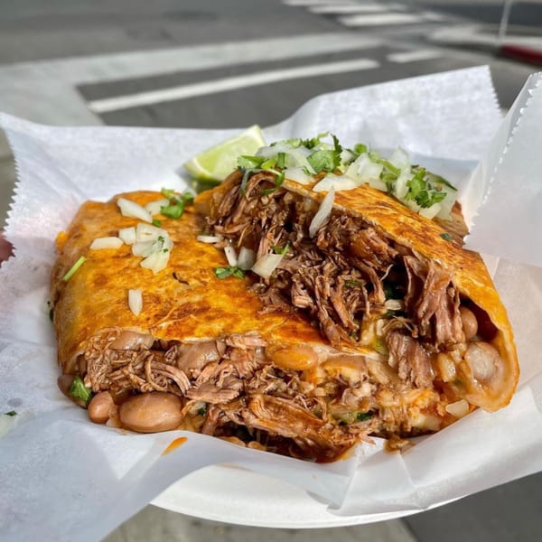 A large, golden-brown birria burrito, overflowing with shredded meat, beans, onions, and cilantro, with a lime wedge.
